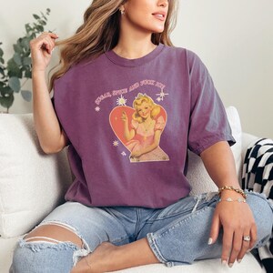 May include: A purple t-shirt with the text "Sugar Spice and Fuck Off" above a graphic of a woman in a pink dress inside a heart. The shirt is worn with blue jeans.