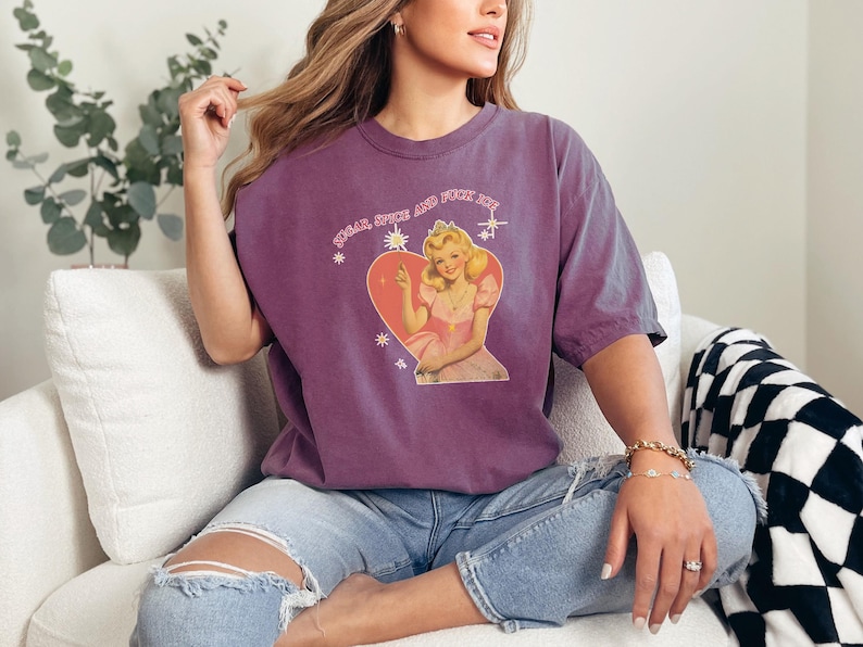 May include: A purple t-shirt with the text "Sugar Spice and Fuck Off" above a graphic of a woman in a pink dress inside a heart. The shirt is worn with blue jeans.