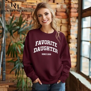 May include: Burgundy crewneck sweatshirt with the text "FAVORITE DAUGHTER" in white block letters, with "SINCE 2000" below. The sweatshirt is part of the Double Mood Collection.