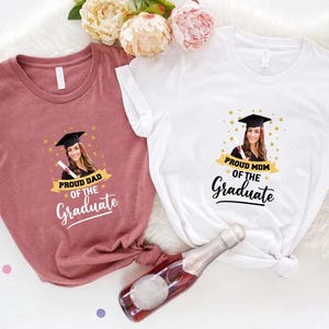 May include: Two t-shirts with a graduation theme. One shirt is pink and says "Proud Dad of the Graduate" with a photo of a graduate in a cap and gown. The other shirt is white and says "Proud Mom of the Graduate" with a photo of a graduate in a cap and gown.