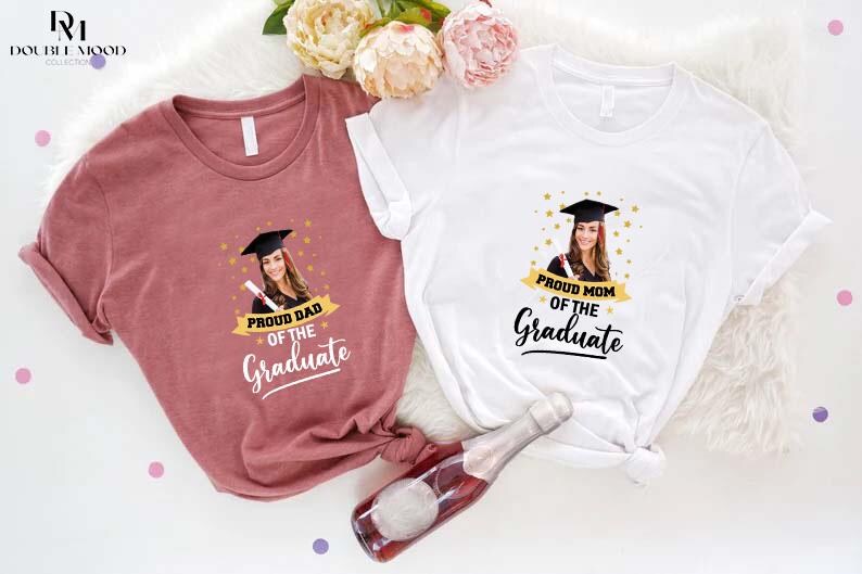 May include: Two t-shirts with a graduation theme. One shirt is pink and says "Proud Dad of the Graduate" with a photo of a graduate in a cap and gown. The other shirt is white and says "Proud Mom of the Graduate" with a photo of a graduate in a cap and gown.