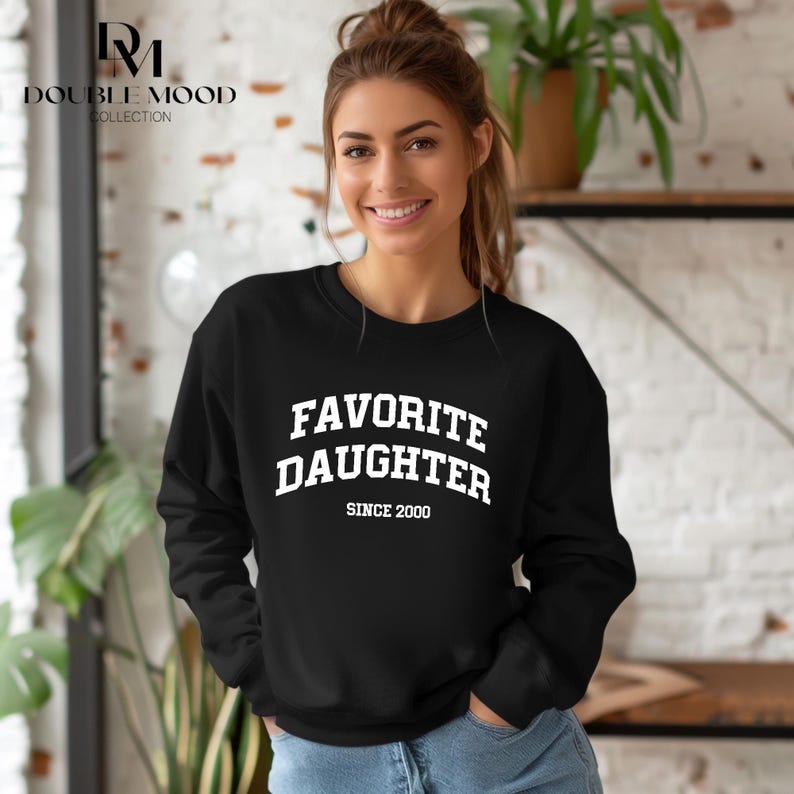 May include: Black sweatshirt with the text "FAVORITE DAUGHTER" in white block letters, with "SINCE 2000" below. The sweatshirt is long-sleeved and has a crew neck.