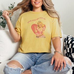 May include: Mustard yellow t-shirt with the text "SUGAR, SPICE AND FUCK ICE" above a vintage-style illustration of a woman in a pink dress inside a pink heart. The shirt is worn with blue jeans.