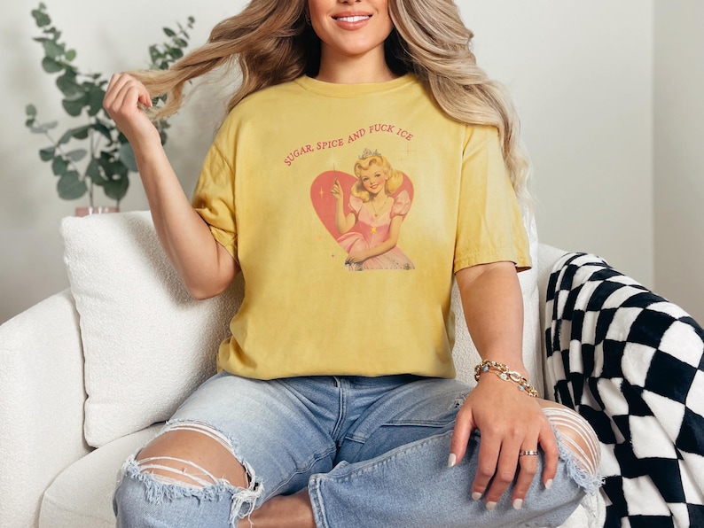 May include: Mustard yellow t-shirt with the text "SUGAR, SPICE AND FUCK ICE" above a vintage-style illustration of a woman in a pink dress inside a pink heart. The shirt is worn with blue jeans.
