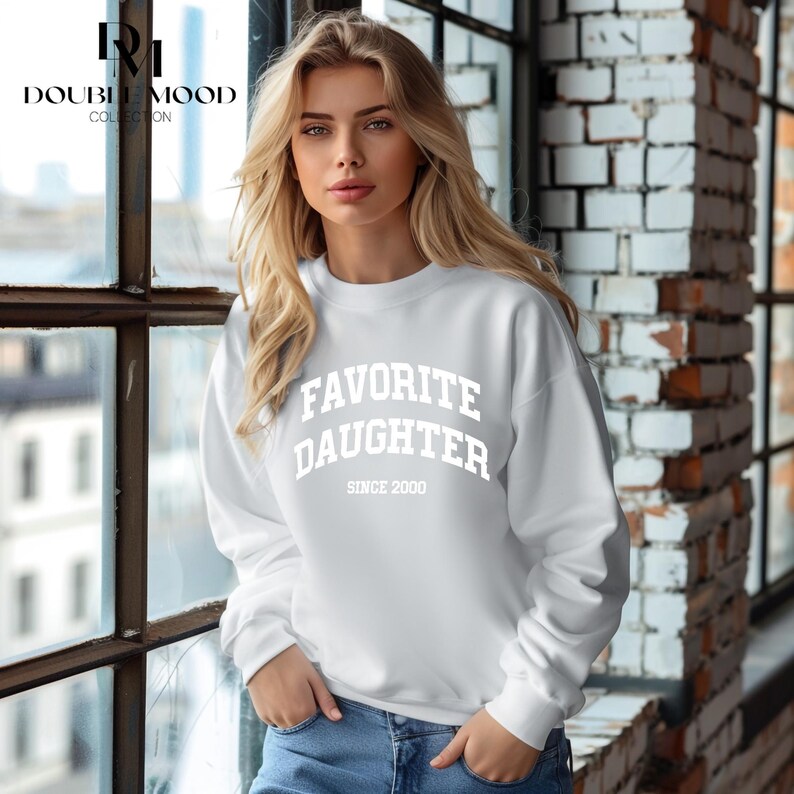 May include: White sweatshirt with the text "FAVORITE DAUGHTER SINCE 2000" in bold, white letters. The sweatshirt is a crew neck style. The model is wearing blue jeans.