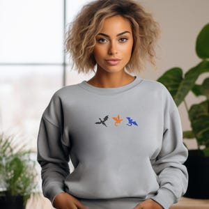 May include: A light gray sweatshirt featuring three embroidered dragon silhouettes in black, orange, and blue. The dragons are arranged horizontally across the chest. The sweatshirt has a crew neck and long sleeves.