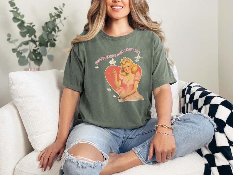 May include: Olive green t-shirt with a graphic of a vintage-style woman in a pink dress inside a heart, with the text "Sugar Spice and Fuck Life". The shirt is worn by a person wearing blue jeans.