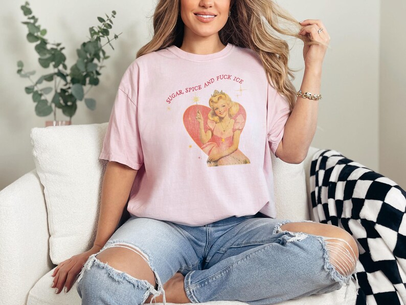 May include: A light pink t-shirt featuring a vintage-style graphic of a woman inside a heart, with the text "SUGAR, SPICE AND FUCK ICE" in a circular arc above the heart. The shirt is worn by a person.