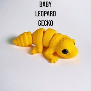 May include: A yellow, segmented, plastic toy gecko with black eyes. The toy is labeled "BABY LEOPARD GECKO".