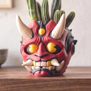 Könnte beinhalten: Ein roter und schwarzer Keramik-Blumentopf in Form einer Oni-Maske mit weißen Hörnern und gelben Augen. Der Topf ist mit grünen Kakteen gefüllt. Der Topf steht auf einer Holzoberfläche.