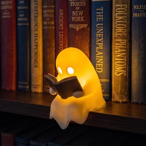 May include: A small, glowing, off-white ghost-shaped lamp made of smooth material, featuring two circular eye cutouts and holding a small, dark gray open book in its hands, positioned on a wooden shelf in front of a row of various hardcover books.