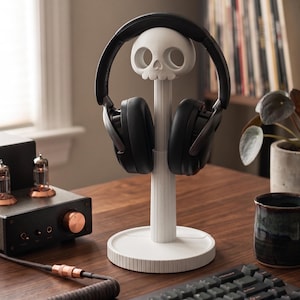 May include: A white skull-shaped headphone stand supports black headphones. The stand is on a wooden desk with a keyboard, a mug, and audio equipment. The skull has eye sockets and a nose cavity.