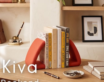 May include: A pair of red bookends, holding several books on a wooden desk. The bookends have a unique triangular design. The word "Kiva" and "Bookend" are printed on the desk. A pen, a small dish with keys, and a stack of notebooks are also on the desk.