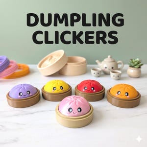 Op de afbeelding: Vijf kleurrijke dumpling-clickers met lachende gezichtjes, elk rustend op een kleine bruine basis. De set bevat paarse, gele, rode, oranje en roze dumplings. De tekst DUMPLING CLICKERS staat erboven. Een houten stoommandje, een kleine theepot, twee kopjes en een vetplantje zijn ook zichtbaar.
