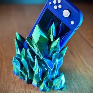 May include: A blue Nintendo Switch Lite video game console is resting in a 3D printed stand made of green and blue crystals.