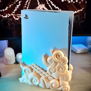 May include: A white PlayStation 5 console with a decorative cloud-shaped stand. The console has a PlayStation logo on the front.
