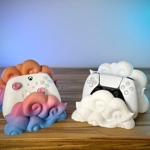 May include: Two controller stands, one for Xbox and one for Playstation, each designed with a cloud motif. The Xbox stand features a white controller with pink accents, set in a cloud with pink, blue, and orange hues.