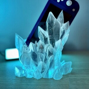 May include: A blue Nintendo Switch console is resting in a clear crystal-shaped stand. The stand is made of translucent plastic and resembles a cluster of ice crystals.