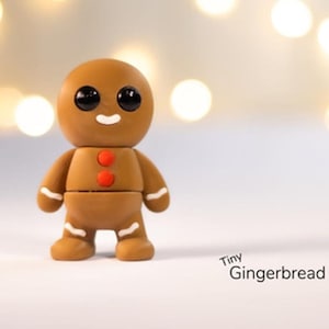 May include: A small gingerbread man figurine with black eyes, a white smile, and red buttons. The figurine is made of brown clay and has a white icing design on its arms and legs. The figurine is standing on a white surface with a blurry background of golden lights.