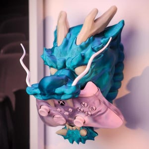 May include: A teal blue 3D printed dragon head wall mount with a pink and white video game controller in its mouth.