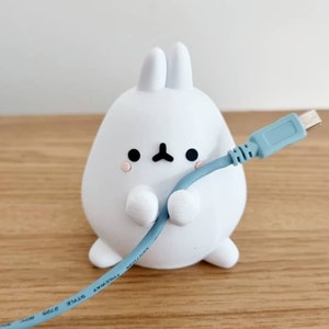 以下が含まれることがあります： 青いUSBケーブルが巻き付けられた、白いウサギの形をしたケーブルホルダー。ケーブルホルダーは木製の表面の上に置かれています。