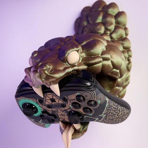 May include: A 3D printed wall mount in the shape of a snake's head, with a textured, metallic green finish. The snake is holding a video game controller in its mouth.