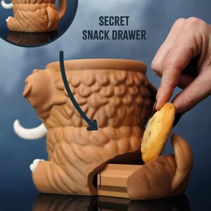 May include: A brown ceramic mammoth-shaped container with a secret snack drawer. The container has a textured surface and white tusks. The text "SECRET SNACK DRAWER" is visible. A hand is placing a cookie into the drawer.