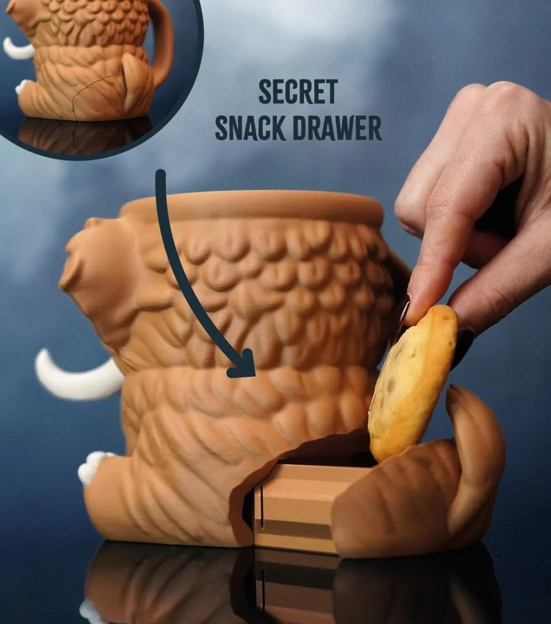 May include: A brown ceramic mammoth-shaped container with a secret snack drawer. The container has a textured surface and white tusks. The text "SECRET SNACK DRAWER" is visible. A hand is placing a cookie into the drawer.