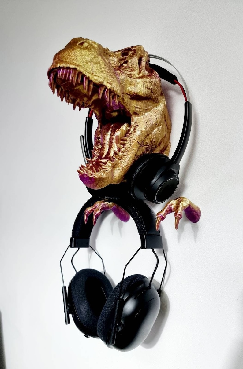May include: A gold-colored Tyrannosaurus Rex head sculpture with pink accents, mounted on a wall and holding a pair of black headphones.