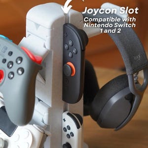 May include: A white 3D-printed gaming accessory with slots for Joy-Cons, controllers, and a headset. The text reads "Joycon Slot Compatible with Nintendo Switch 1 and 2". The accessory holds a black Joy-Con, a gray controller, and a black headset.