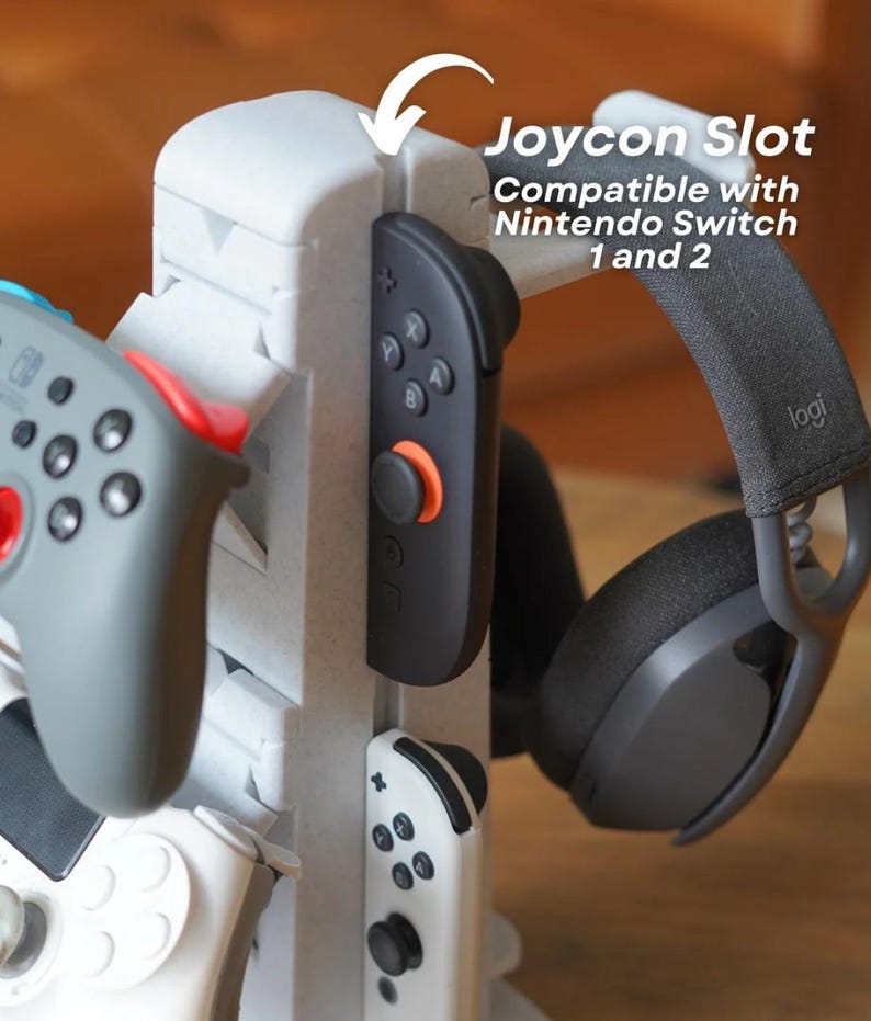 May include: A white 3D-printed gaming accessory with slots for Joy-Cons, controllers, and a headset. The text reads "Joycon Slot Compatible with Nintendo Switch 1 and 2". The accessory holds a black Joy-Con, a gray controller, and a black headset.