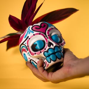 May include: A Day of the Dead skull-shaped planter, white with blue, pink, and black decorative accents. The skull has large blue eye sockets and a blue mouth. The planter holds a plant with dark red leaves, set against a yellow background.