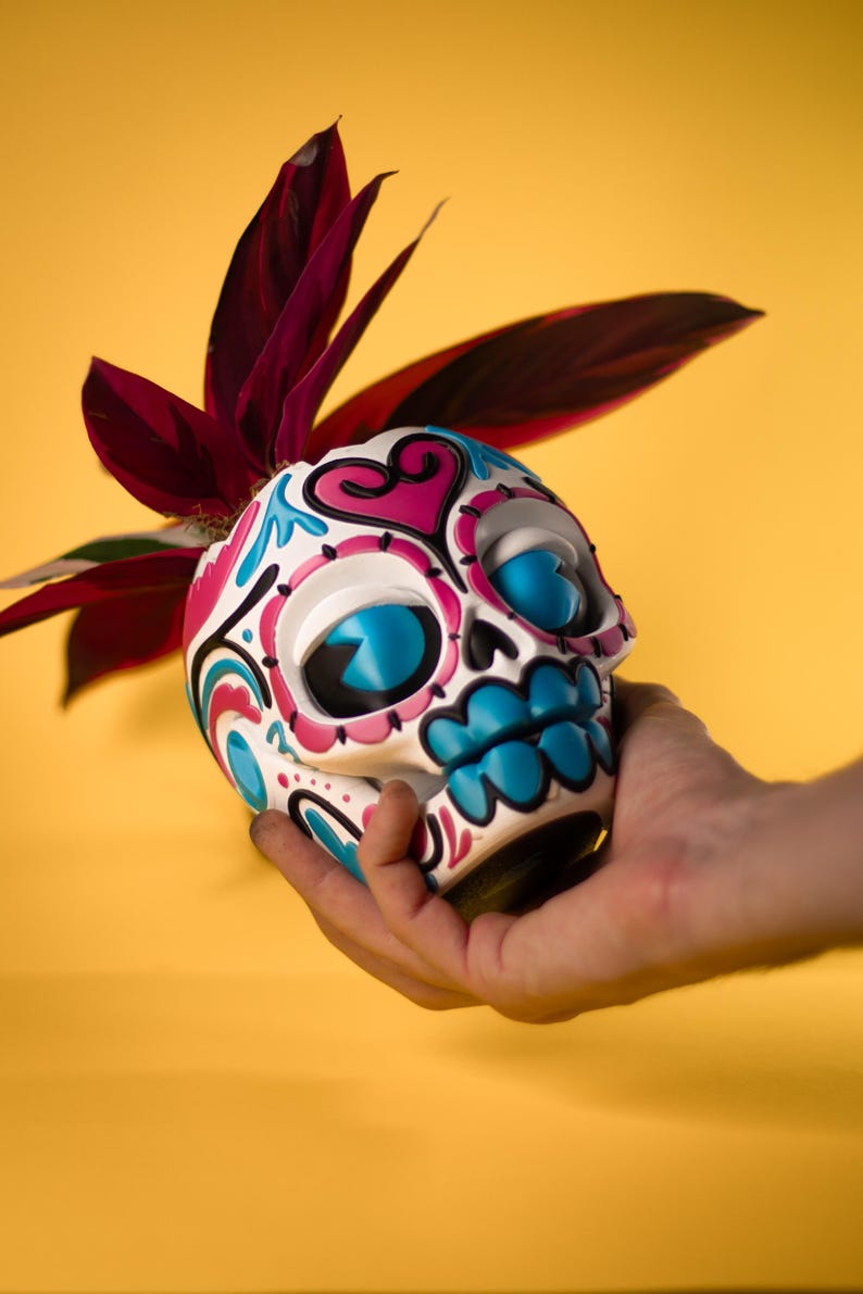 May include: A Day of the Dead skull-shaped planter, white with blue, pink, and black decorative accents. The skull has large blue eye sockets and a blue mouth. The planter holds a plant with dark red leaves, set against a yellow background.