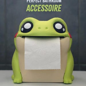 May include: A whimsical frog-shaped toilet paper holder in shades of green and beige. The frog has large, expressive eyes and red cheek accents. The text "PERFECT BATHROOM ACCESSOIRE" is displayed above the frog. The holder dispenses white toilet paper.