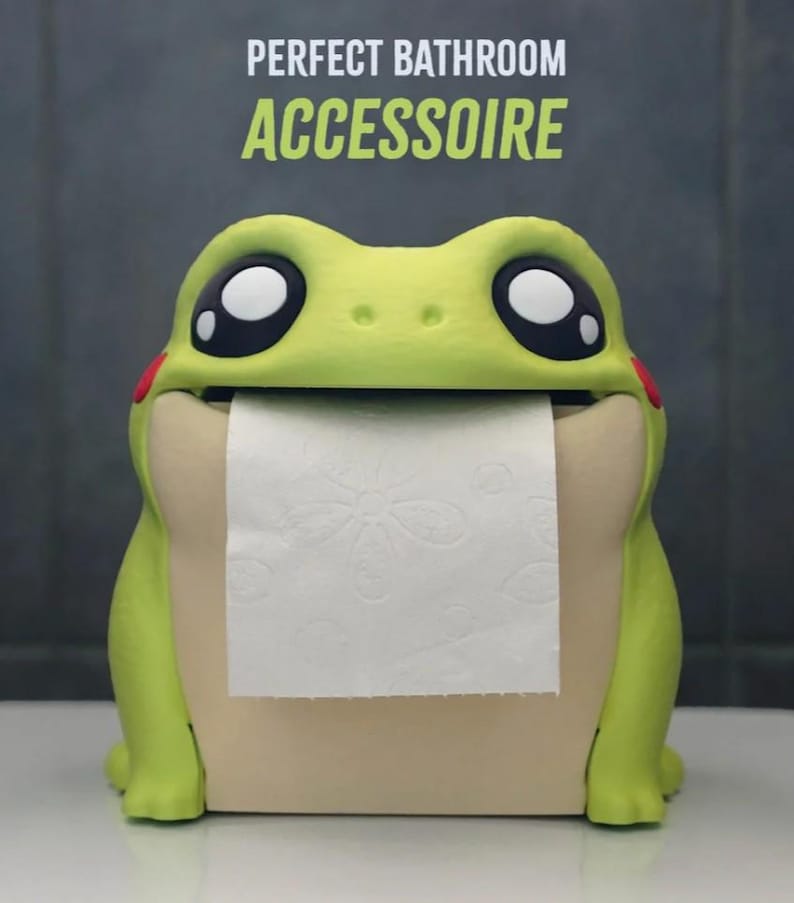 May include: A whimsical frog-shaped toilet paper holder in shades of green and beige. The frog has large, expressive eyes and red cheek accents. The text "PERFECT BATHROOM ACCESSOIRE" is displayed above the frog. The holder dispenses white toilet paper.