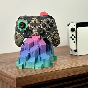 May include: A black and green video game controller with a gold and black design sits on a colorful crystal-shaped stand. A white Nintendo Switch console is in the background.