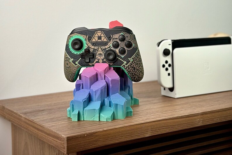 May include: A black and green video game controller with a gold and black design sits on a colorful crystal-shaped stand. A white Nintendo Switch console is in the background.