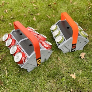 May include: Two gray and black beverage carriers with red handles. Each carrier holds eight cans and has the text "8 Pack" printed on the side. The carriers are sitting on a green grassy surface.