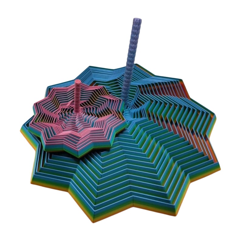 May include: A colorful, abstract sculpture featuring layered, star-shaped forms in shades of blue, green, pink, and orange. Two pink and blue cylindrical elements rise from the structure. The artwork has a geometric, modern aesthetic.