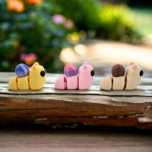 May include: Three small, colorful, clay figurines of snails. The snails are yellow, pink, and beige with purple, pink, and brown shells. The snails are arranged in a row on a wooden surface.