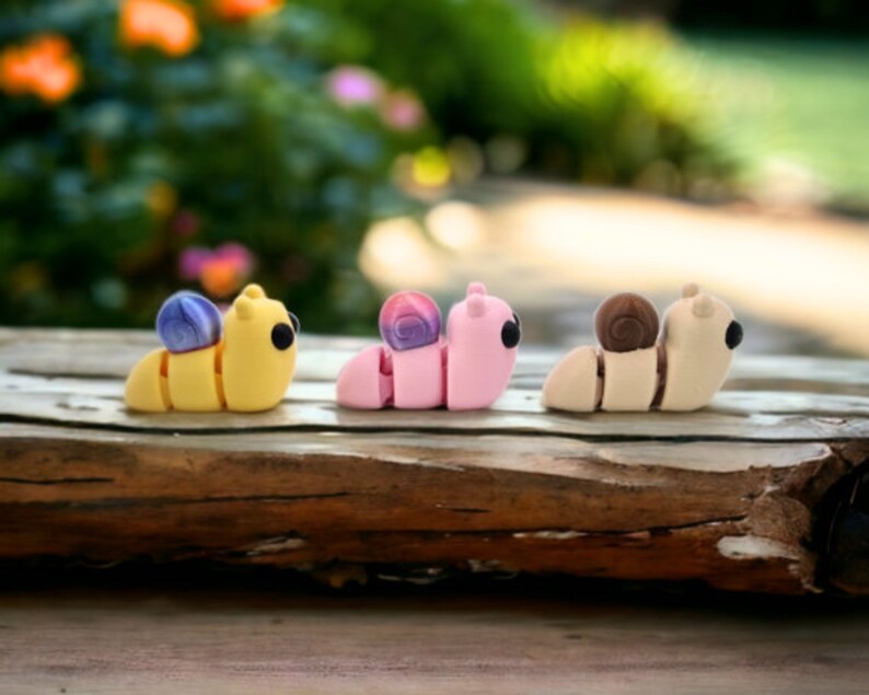 May include: Three small, colorful, clay figurines of snails. The snails are yellow, pink, and beige with purple, pink, and brown shells. The snails are arranged in a row on a wooden surface.