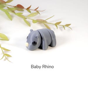 K&ouml;nnte beinhalten: Eine kleine graue Baby-Nashorn-Figur mit schwarzen Augen und einem hellbraunen Horn. Die Figur liegt auf dem Bauch mit den Beinen unter sich. Die Figur befindet sich auf einem wei&szlig;en Hintergrund.