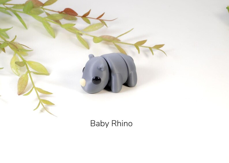 K&ouml;nnte beinhalten: Eine kleine graue Baby-Nashorn-Figur mit schwarzen Augen und einem hellbraunen Horn. Die Figur liegt auf dem Bauch mit den Beinen unter sich. Die Figur befindet sich auf einem wei&szlig;en Hintergrund.