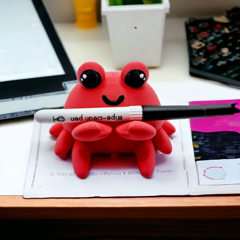 May include: A red 3D printed crab with black eyes and a smile holds a white dry-erase marker with the text "Wipe-clean pen" in black. The crab is sitting on a white surface.