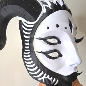 May include: A white and black mask with a stylized design. The mask features large black horns, black accents around the eyes and mouth, and a series of black lines along the sides. The eyes are white with black outlines. The mask has a smooth, matte finish.