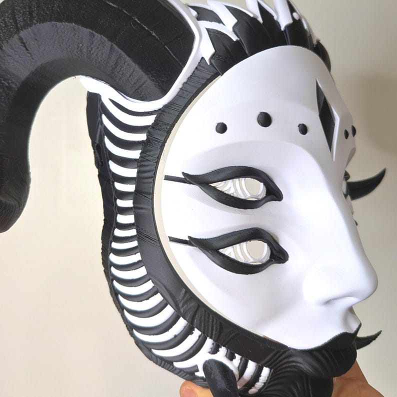 May include: A white and black mask with a stylized design. The mask features large black horns, black accents around the eyes and mouth, and a series of black lines along the sides. The eyes are white with black outlines. The mask has a smooth, matte finish.
