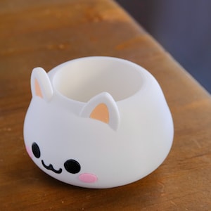 May include: White ceramic planter shaped like a cartoon bunny with pink cheeks and black eyes. The planter has a round opening at the top.