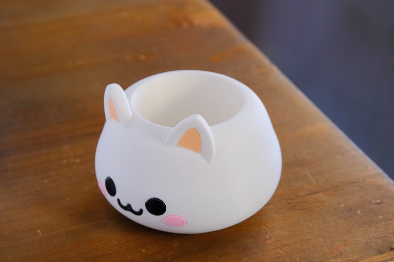 May include: White ceramic planter shaped like a cartoon bunny with pink cheeks and black eyes. The planter has a round opening at the top.