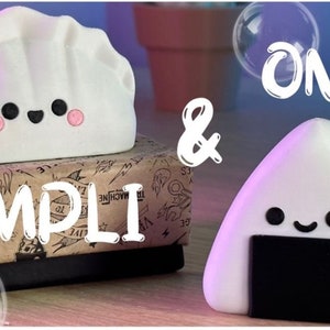 May include: White and black cartoon characters shaped like a dumpling and a rice ball. The dumpling has a pink blush and the rice ball has a black seaweed wrapper. The text "ONI & DUMPLI" is written in white on a brown background.