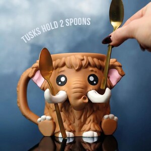 May include: A brown ceramic mug shaped like a cartoon mammoth with a trunk, tusks, and furry details. The mug has a handle and holds two gold spoons. Text on the image reads "TUSKS HOLD 2 SPOONS".